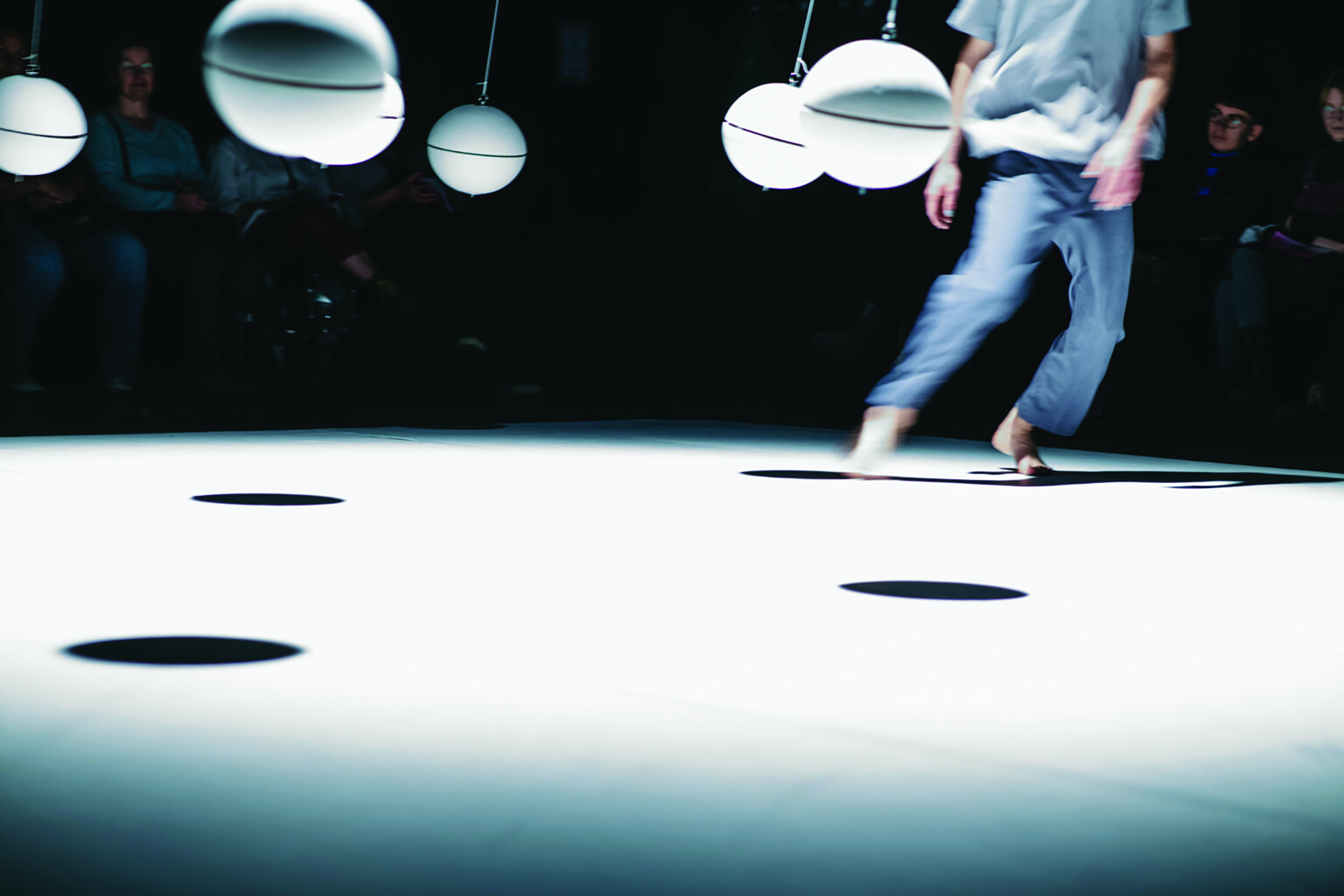 Un artiste vêtu de blanc danse sur un sol blanc devant un public assombri et des boules blanches suspendues par des fils