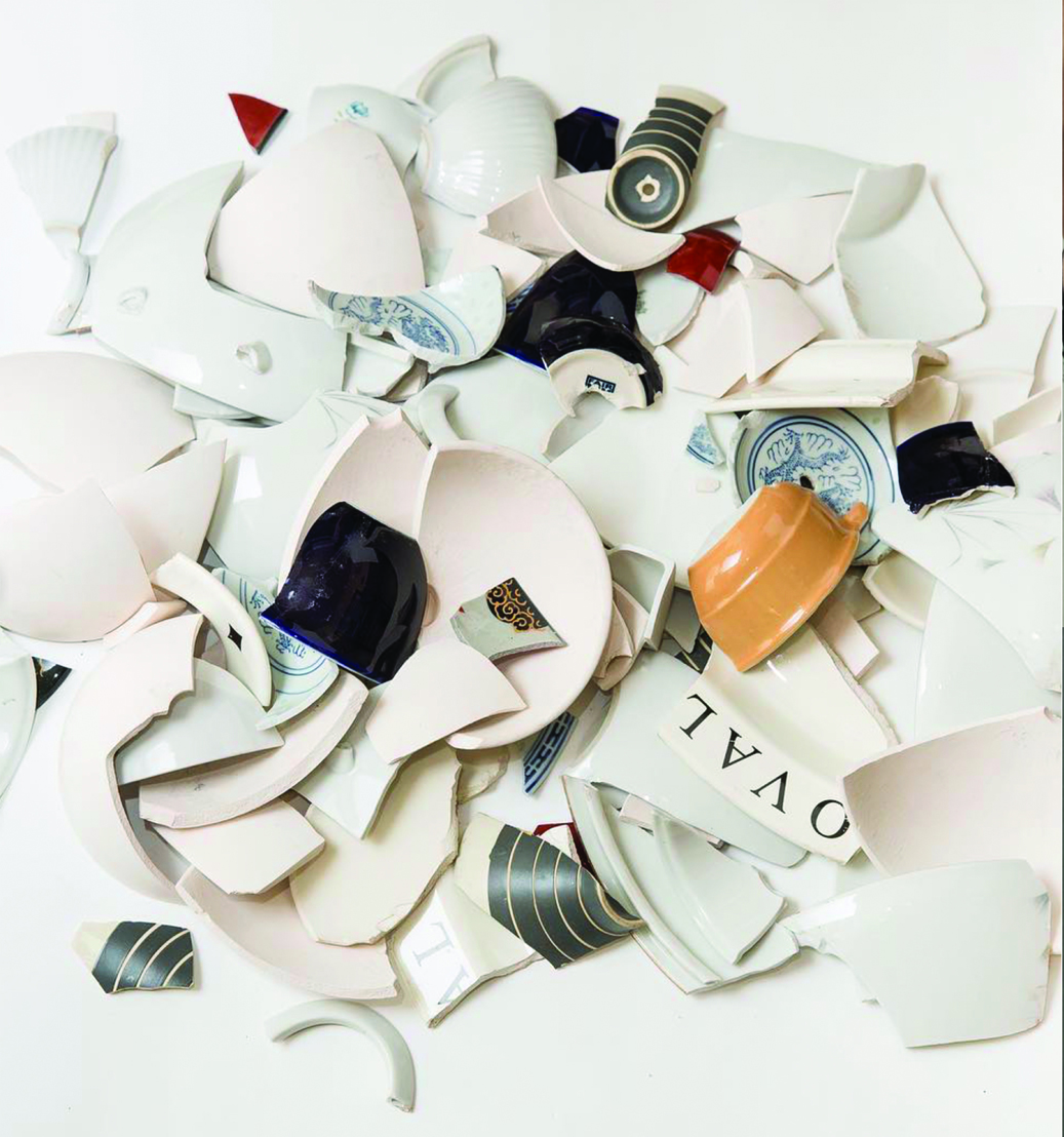 De la vaisselle cassée est disposée sur fond blanc ; des débris blancs et quelques morceaux gris, bleus, beiges et rouges