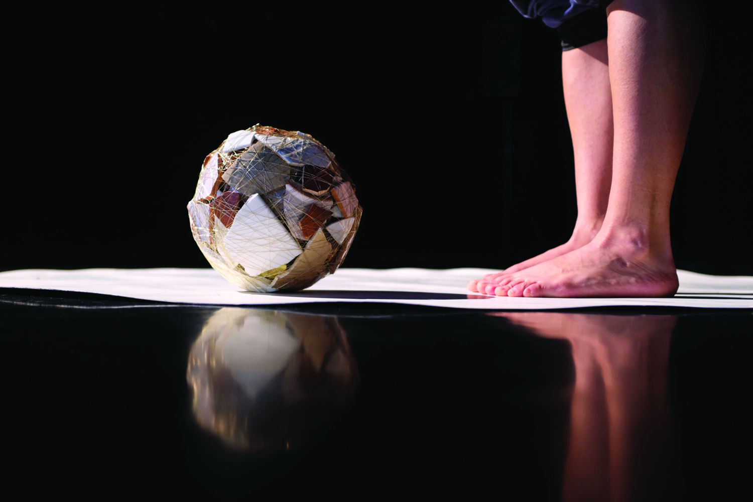 Sur fond noir, des morceaux de vaisselle ficelés forment une boule posée sur un tapis blanc face aux pieds nus d’un artiste