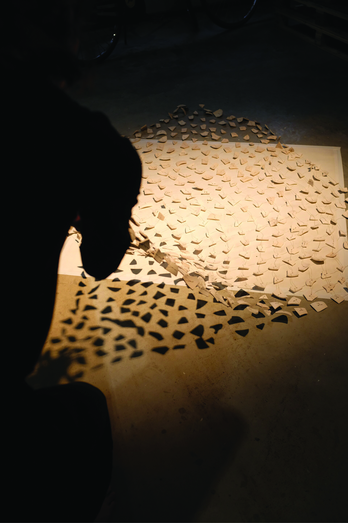 Une silhouette accroupie tient un ensemble de pièces beiges reliées en quadrillage par des fils et disposées sur un sol blanc