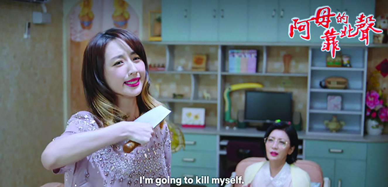 Ruo-min is pictured holding a cleaver to her neck while a distressed Ru-rong watches in the background. The subtitles in English for this Minnan portion of the episode read, “I’m going to kill myself.” The top right-hand corner of the image features the bright-red lettering of this fictional drama’s title, “The Sound of Mother’s Lament” (阿母的靠北聲).
