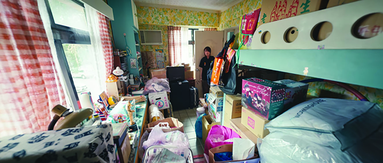Ruo-min is pictured standing at the doorway of her own room. Her bedroom resembles a storage facility, with boxes and packages stacked on her bed and around her desk.