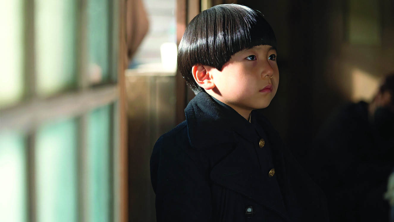 Pachinko’s TV screenshot of Noa, a child character performed by actor Park Jae-joon. He is wearing an elementary school uniform and standing in front of the window and looks very tense and upset. This screenshot is from episode 8.