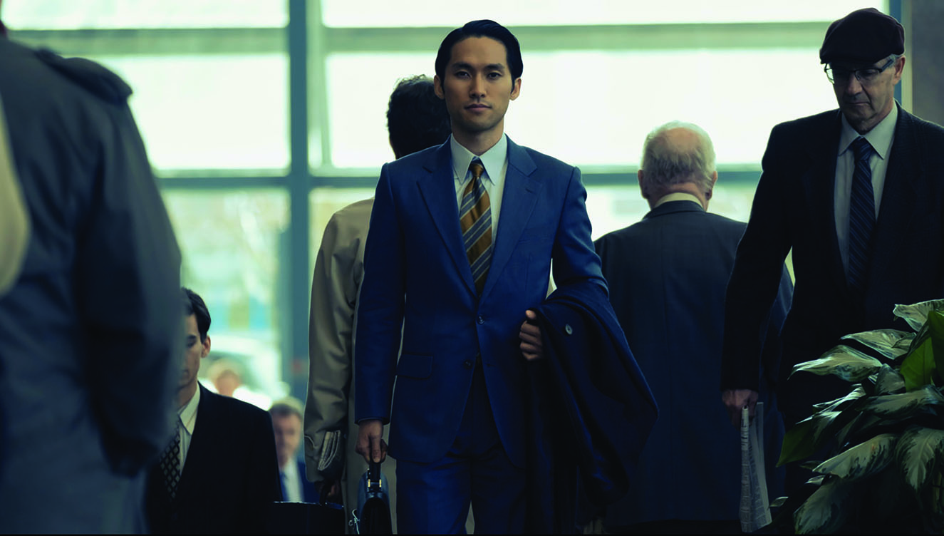 Pachinko’s TV screenshot of Solomon, a character performed by actor Jin Ha. He is walking in a modern building at Wall Street among the crowd, who are predominately white. He is dressed in professional suit and carrying a briefcase and a coat.