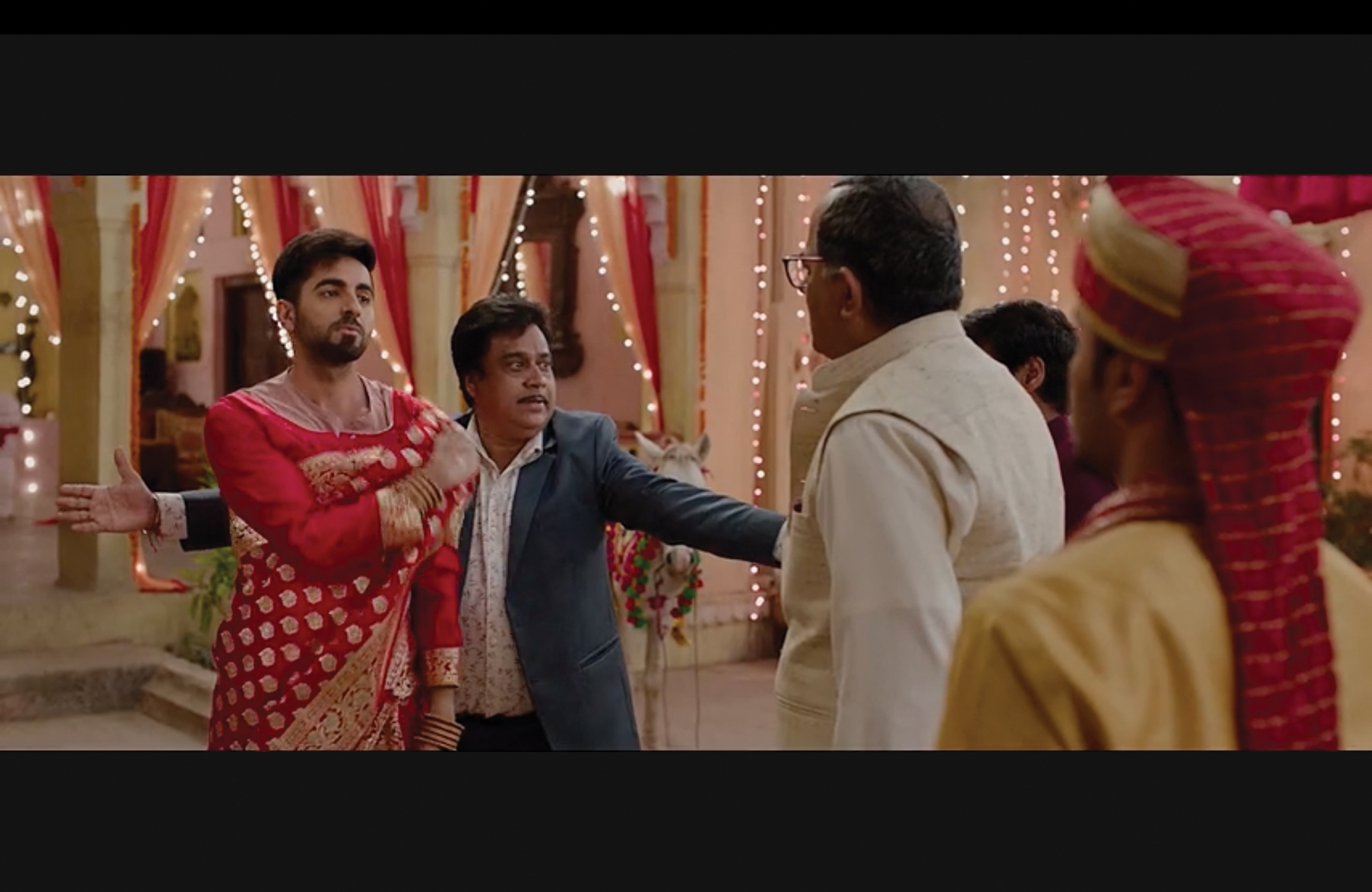 Kartik stands defiantly in front of his father as another man stands behind Kartik as if to protect him from confrontation.