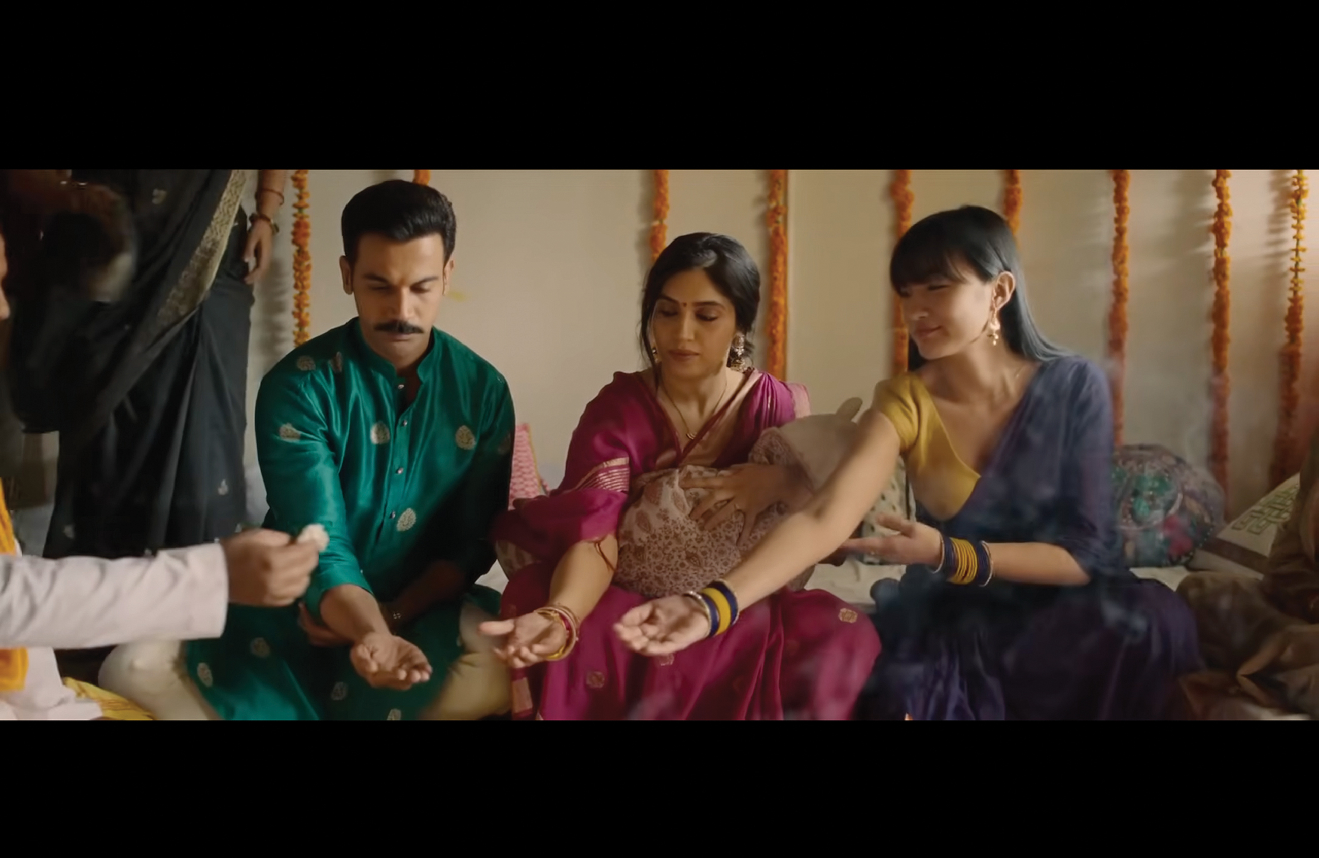 A man and two women sit in a row with their hands held out. All three wear traditional ceremony clothing.